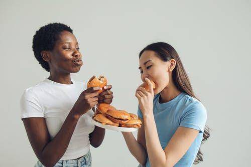Women in white and blue shirts eating burgers (pexels/Polina Tankilevitch)