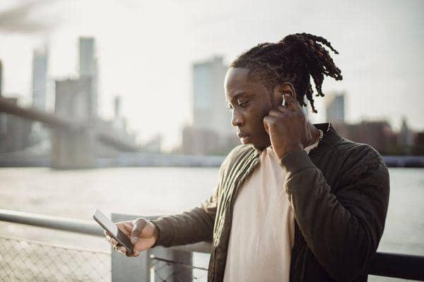 Seorang pria berambut dreadlocks mengenakan earphone sambil melihat ponsel di tepi sungai dengan latar gedung kota.