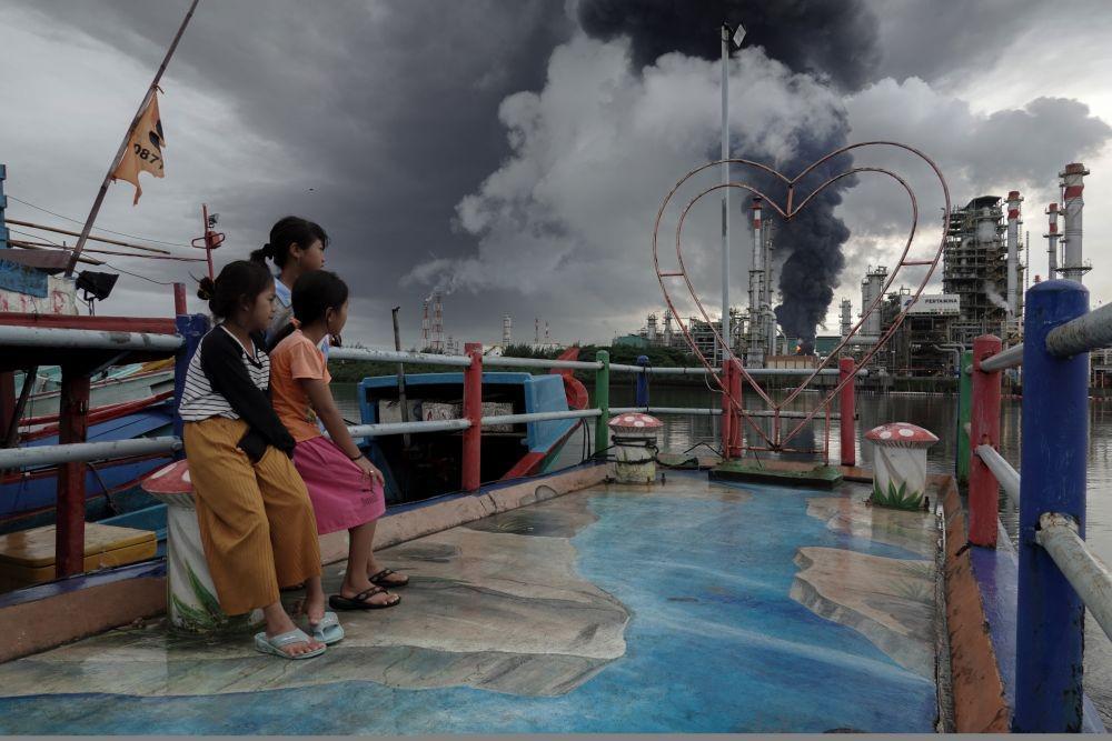Beberapa anak melihat kepulan asap hitam dari tangki 36 T 102 yang terbakar di Kilang Pertamina Internasional RU IV Cilacap, Jawa Tengah, Minggu (14/11/2021). Upaya pemadaman masih terus dilakukan dengan melakukan penyemprotan untuk mengisolir tangki yang terbakar agar api tidak merambat ke tangki yang lain. ANTARA FOTO/Idhad Zakaria