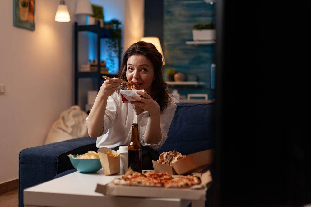 Seorang wanita sedang makan sambil nonton TV di malam hari (Shutterstock/DC Studio)