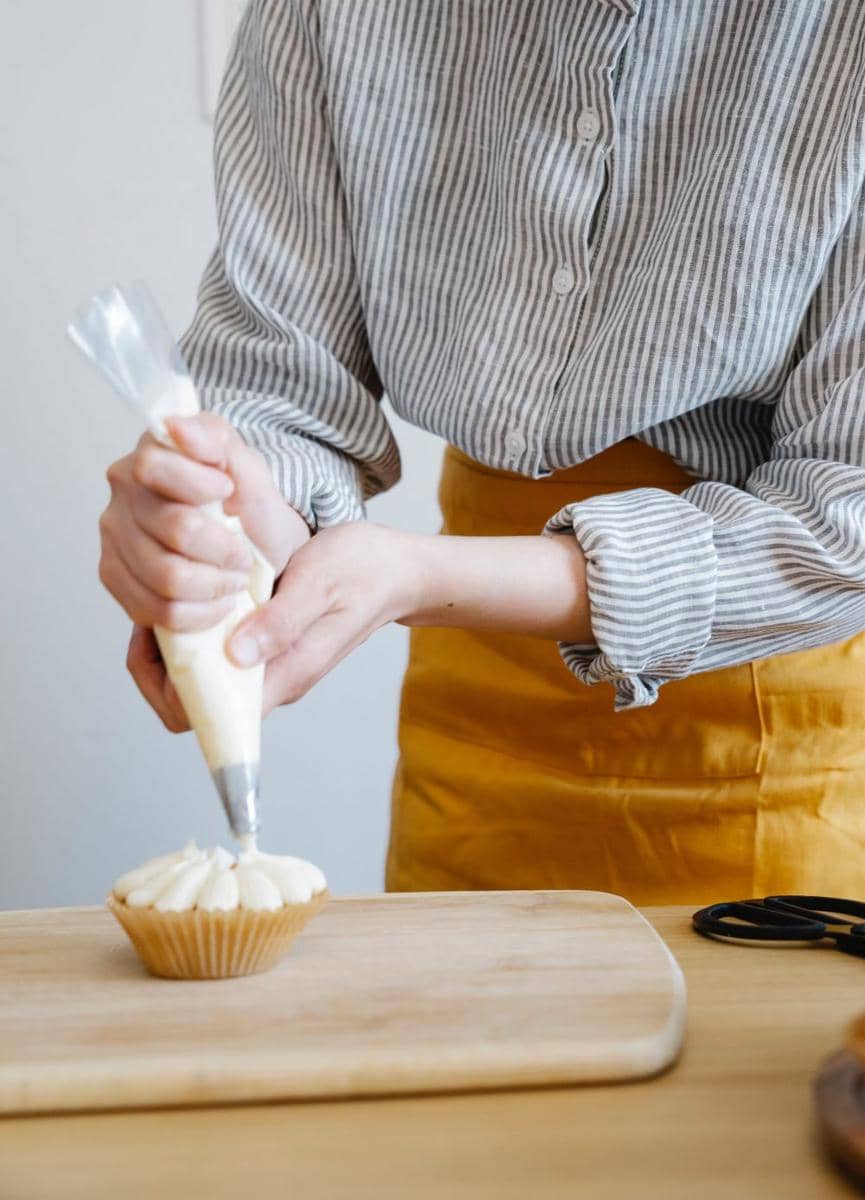 Tangan menghias kue bundar berlapis frosting putih dengan whipped cream menggunakan spatula di atas meja dapur.