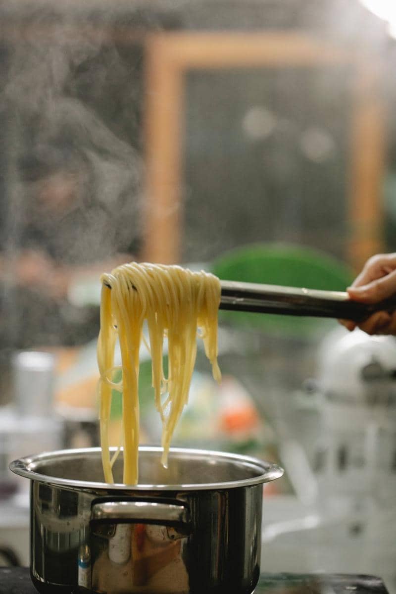 Seorang pria berdiri di dapur sambil mengaduk mi yang sedang direbus dalam panci di atas kompor gas.
