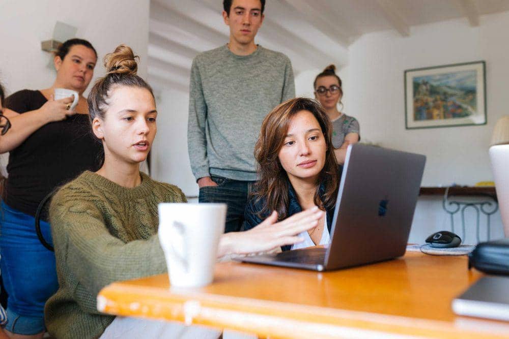 Seorang perempuan memimpin rapat bersama beberapa rekan kerja yang duduk mengelilingi meja di ruang kantor modern.