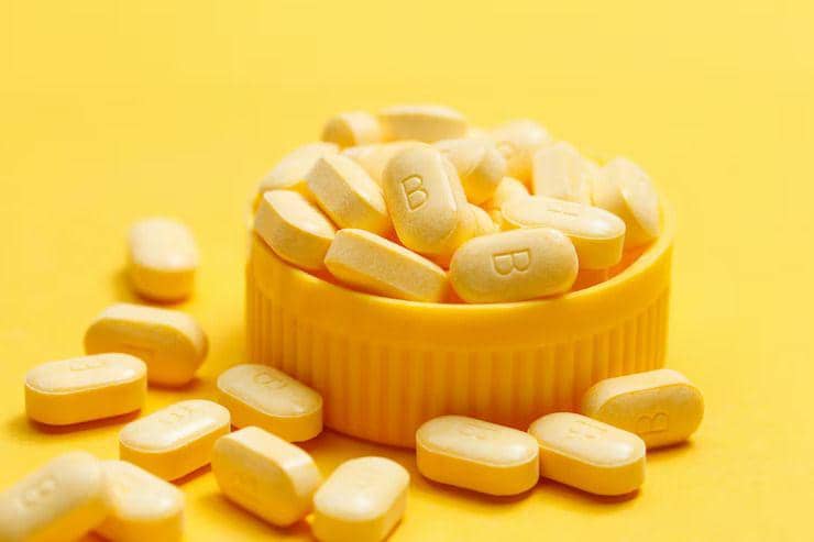 Beberapa tablet suplemen biotin berwarna kuning dengan huruf B di atasnya tersusun di atas tutup wadah berwarna senada.