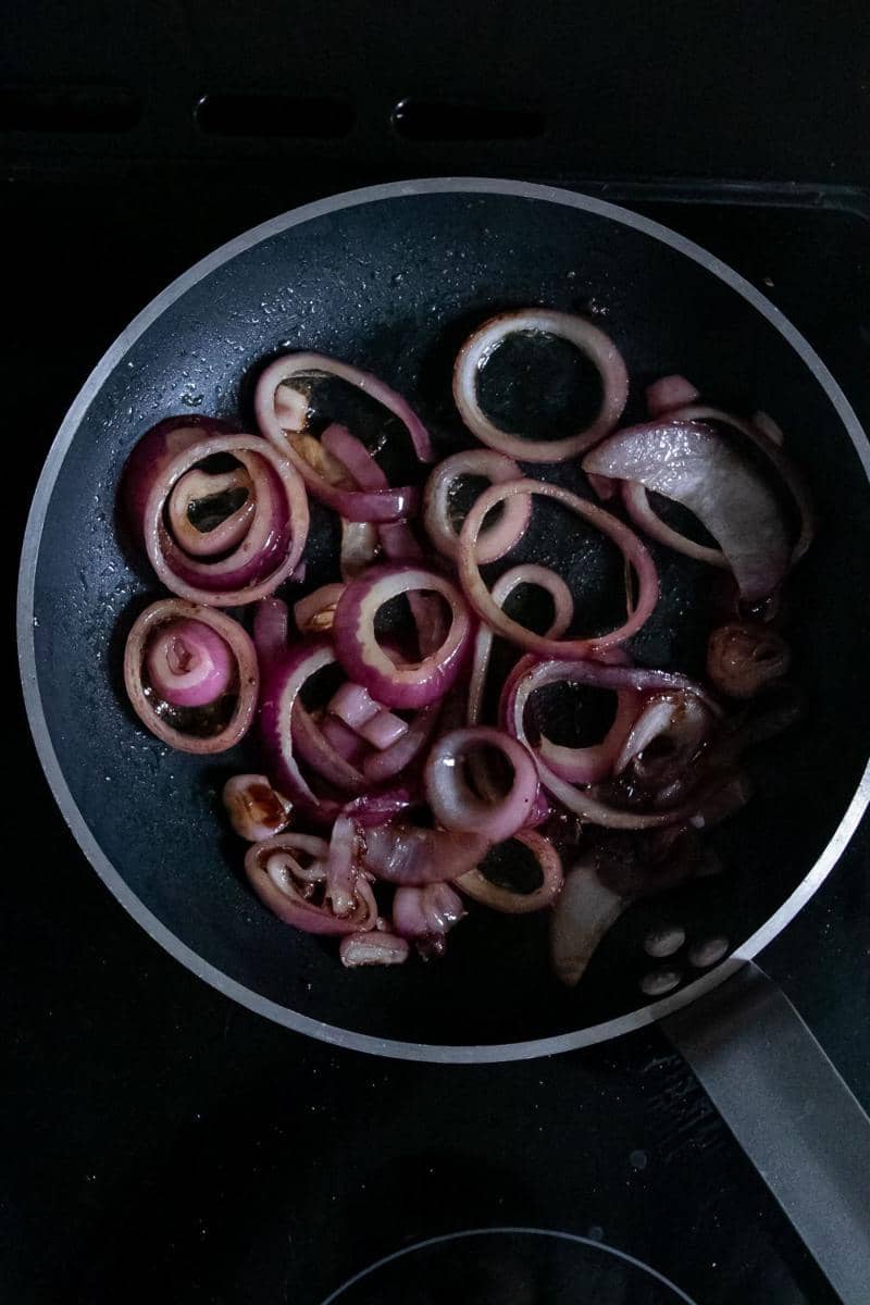 Tangan menumis irisan bawang di wajan dengan spatula kayu, ilustrasi proses memasak di dapur rumah.