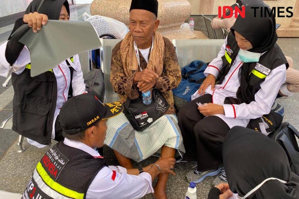 Petugas haji membantu seorang jemaah lansia yang duduk di kursi sambil memeriksa kakinya di area luar ruangan saat pelaksanaan ibadah haji.