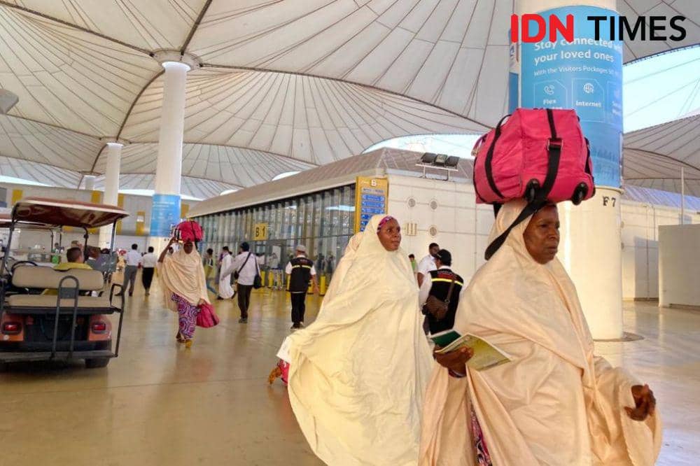 Jemaah haji asal Afrika mengenakan pakaian ihram berjalan di area kedatangan Bandara Jeddah dengan membawa tas besar berwarna merah muda.
