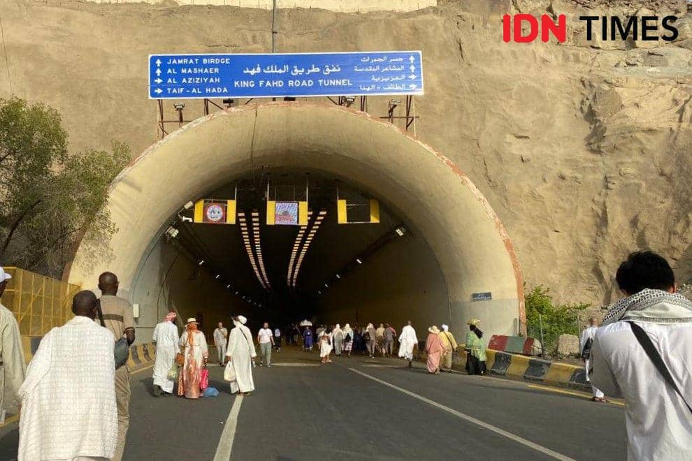 Jemaah haji berjalan menuju terowongan King Fahd Road di Mina, Arab Saudi, dengan latar pegunungan dan papan petunjuk arah berbahasa Arab dan Inggris.