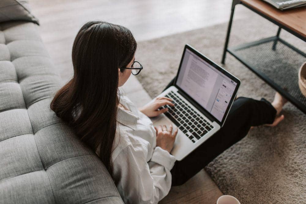 Seorang perempuan mengenakan sweater kuning mengetik di laptop sambil duduk di meja kerja dengan minuman.