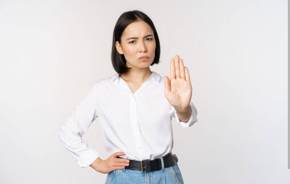 Seorang wanita berambut pendek mengenakan kemeja putih dan celana jeans mengangkat tangan dengan telapak terbuka sebagai isyarat menolak.