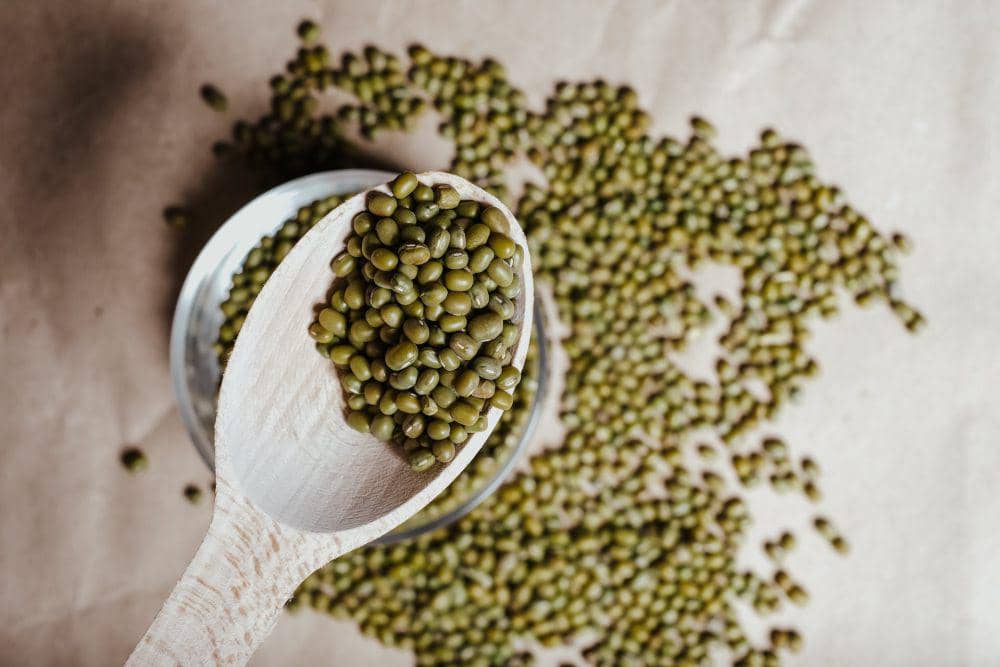 Semangkuk kacang hijau kering berwarna hijau dengan sendok kayu, dikelilingi biji yang tersebar di atas latar putih.