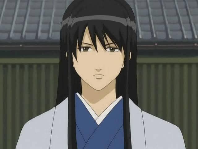 Katsura Kotarou, karakter anime berambut panjang hitam mengenakan kimono biru dan jubah abu-abu, berdiri di depan bangunan tradisional Jepang.