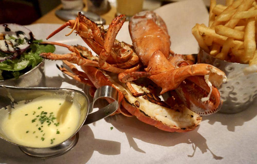 Seekor lobster panggang berwarna merah disajikan di atas piring dengan irisan lemon dan bumbu hijau.