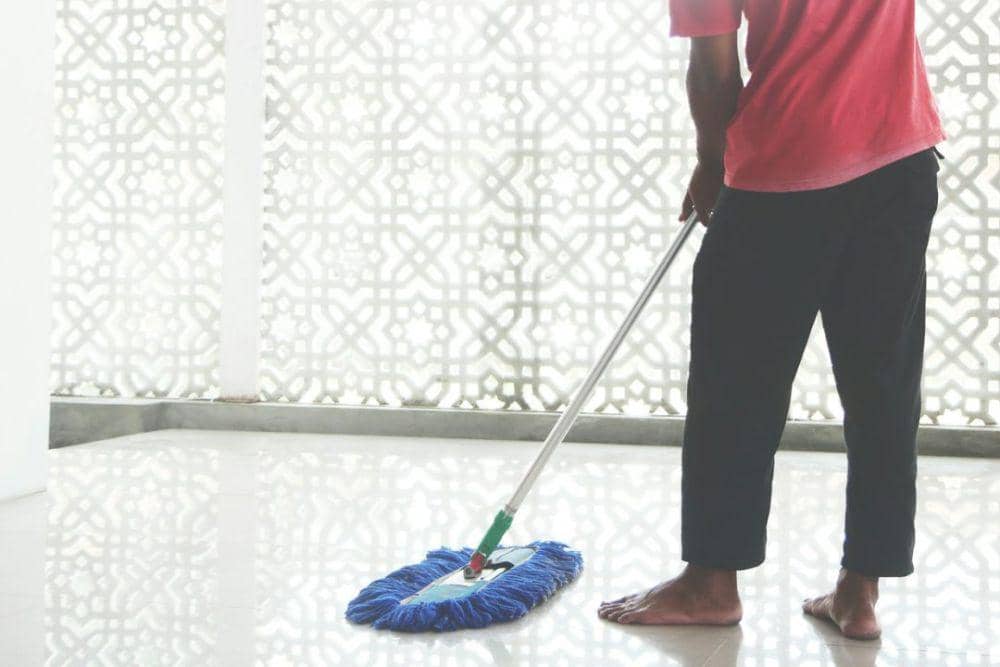 Seseorang mengenakan kaus merah dan celana hitam sedang mengepel lantai masjid dengan alat pel lantai berwarna biru.