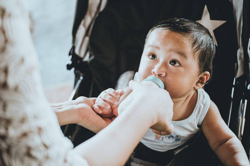 Seorang bayi berbaring sambil minum susu dari botol yang dipegang orang dewasa dengan latar berwarna terang.