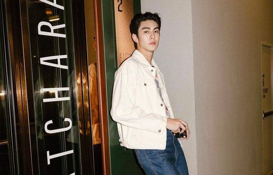 Seorang pria mengenakan jaket denim putih dan celana jeans berdiri di dekat pintu kaca dengan tulisan besar di dinding.