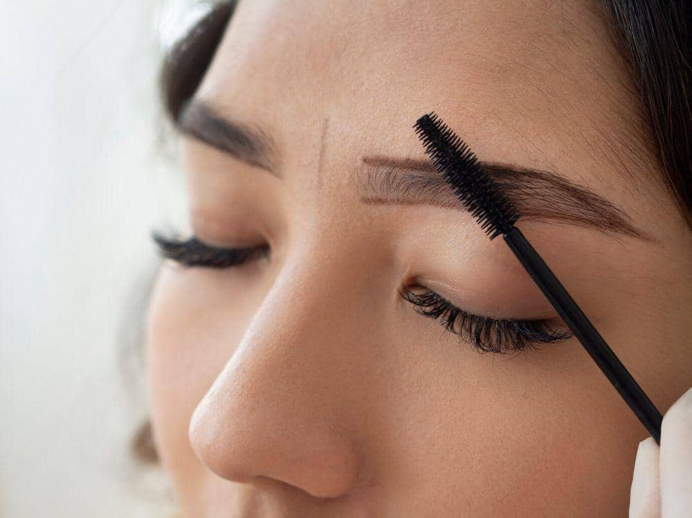 Seorang wanita sedang merapikan alis menggunakan spoolie brush hitam dengan mata tertutup, menampilkan detail riasan alis alami.