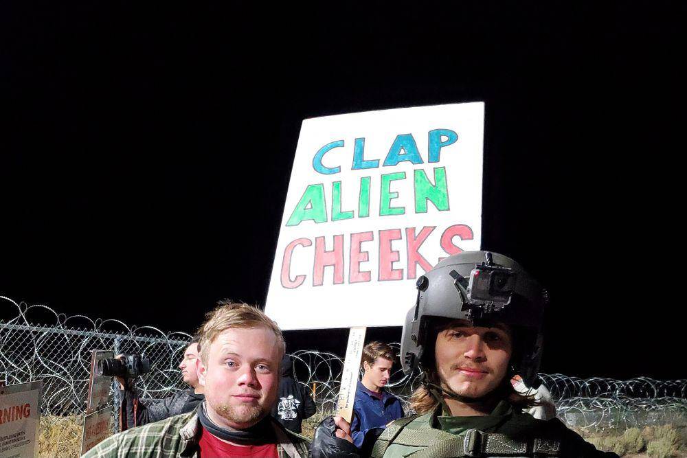 Seorang pengunjuk rasa di Area 51 yang mengenakan baju tempur membawa plakat protes yang bertuliskan, "Tepuk Pipi Alien." (commons.wikimedia.org/David James Henry)