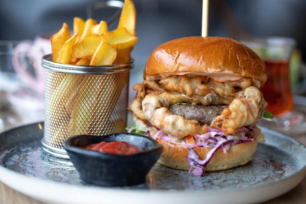 Burger dengan isian daging, sayuran, dan bawang goreng disajikan bersama kentang goreng dan saus tomat di atas piring logam.