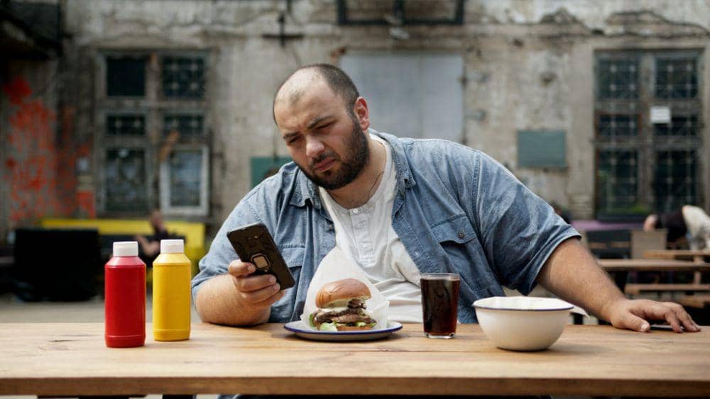 Seorang pria duduk di meja kayu sambil memegang ponsel dan memakan burger dengan minuman soda di depannya di area luar ruangan.
