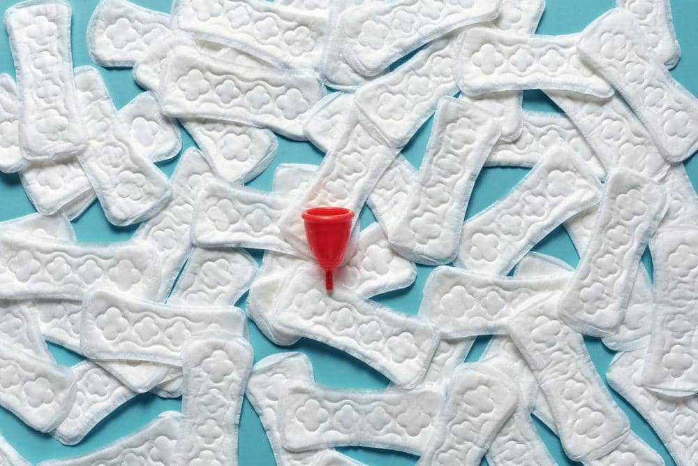 Beberapa pembalut berwarna putih tersusun di atas permukaan biru dengan satu menstrual cup merah di tengahnya.