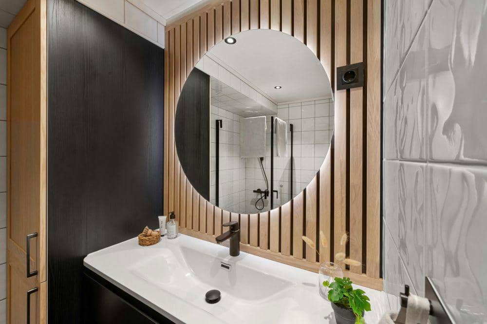 Kamar mandi modern dengan wastafel putih, cermin bundar besar, dan dinding kayu vertikal berpadu ubin putih minimalis.