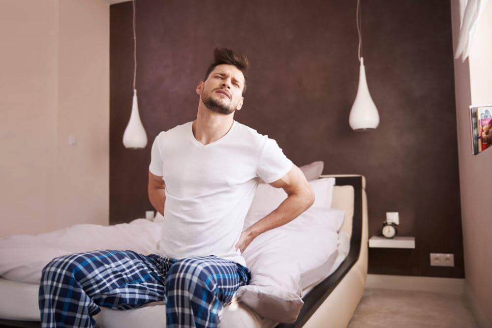 Seorang pria duduk di tepi tempat tidur sambil memegang punggung bagian bawah dan menunjukkan ekspresi kesakitan di kamar tidur.