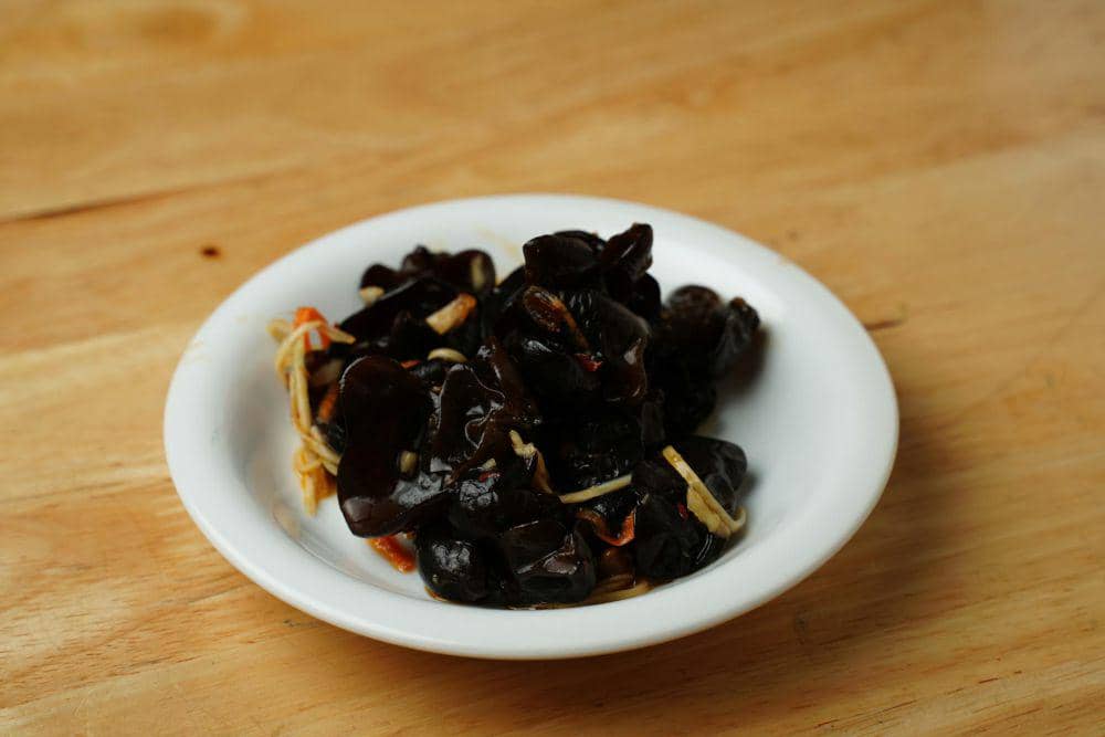 Sekelompok jamur kuping berwarna cokelat tumbuh bertumpuk pada batang kayu, dengan latar belakang hijau samar.