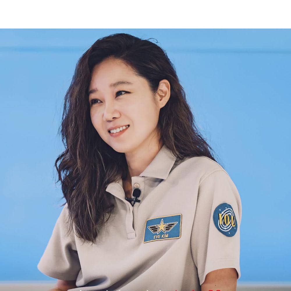 Seorang wanita berambut panjang bergelombang mengenakan seragam abu-abu dengan nama tag Eve Kim dan latar belakang biru cerah.