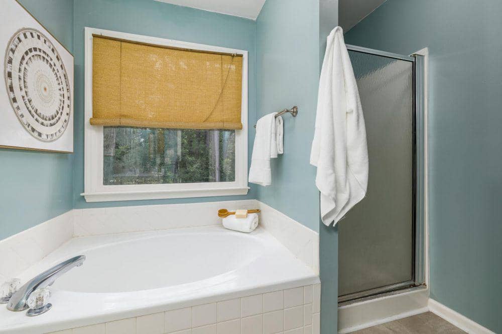 Kamar mandi dengan dinding biru muda, bathtub putih di dekat jendela berpenutup bambu, serta area shower tertutup kaca buram.