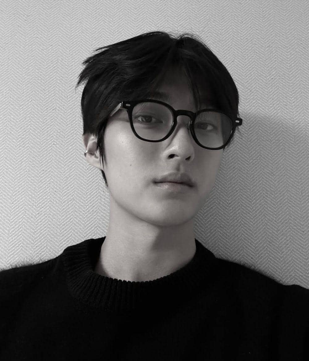 Seorang pria berambut hitam mengenakan kacamata bulat dan sweter gelap berpose di depan dinding bermotif sederhana.