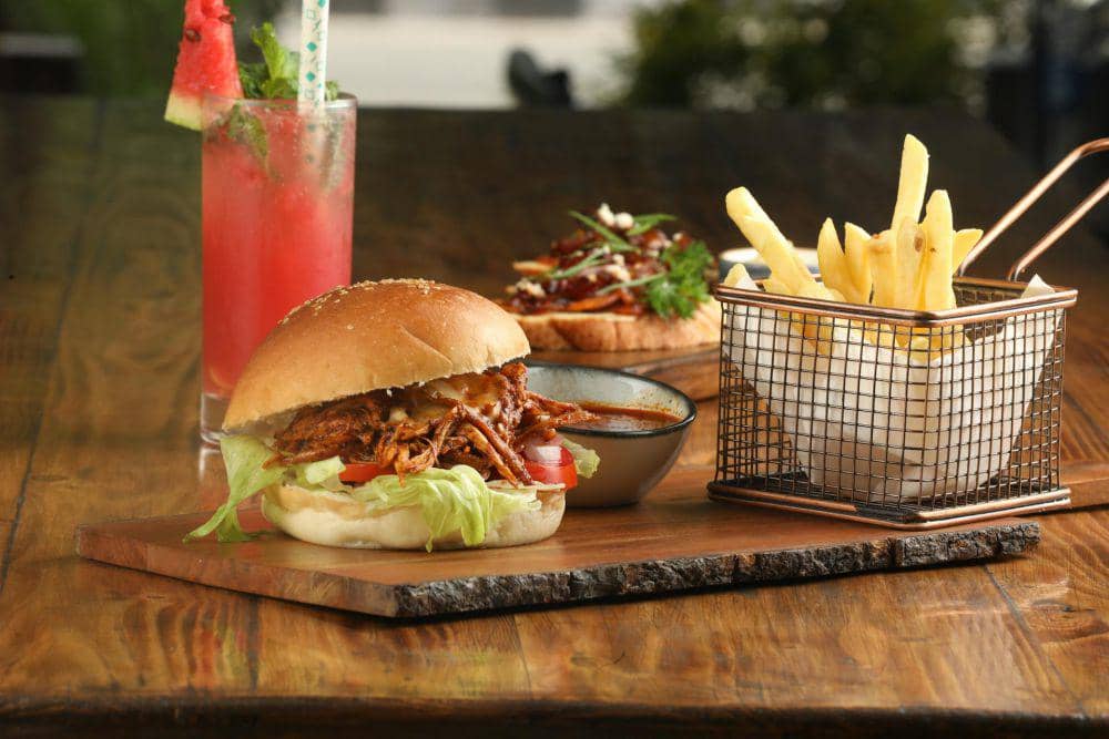 Burger dengan daging suwir, selada, dan tomat disajikan bersama kentang goreng, saus, dan minuman semangka di atas meja kayu.