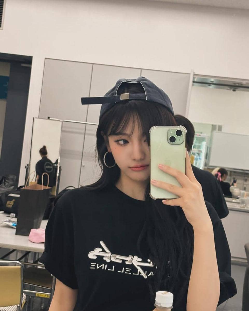Seorang perempuan berambut panjang mengenakan kaus hitam dan topi terbalik berfoto selfie di depan cermin dengan ponsel hijau.