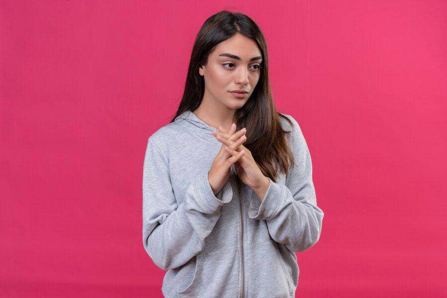 Perempuan berambut panjang mengenakan hoodie abu-abu berdiri dengan ekspresi sedih dan tangan saling menggenggam di latar belakang merah muda.