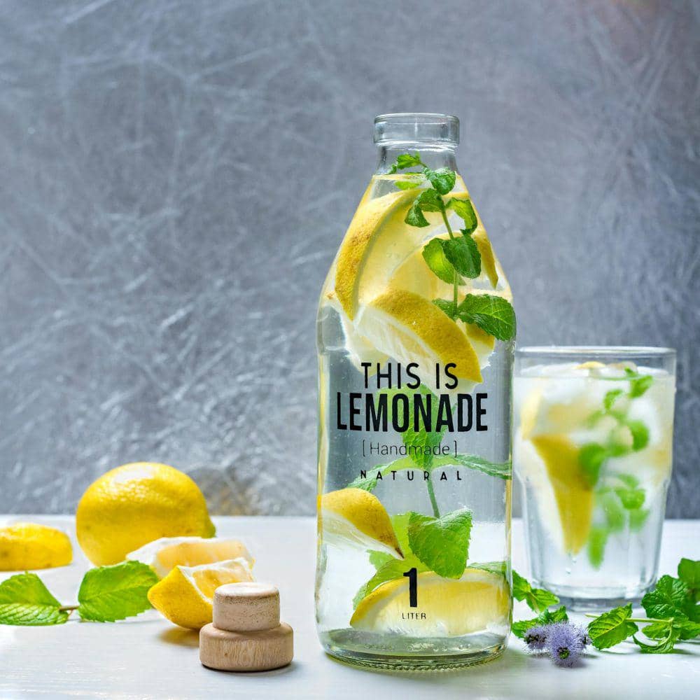 Botol berisi air lemon dengan irisan lemon dan daun mint segar di atas meja, disertai gelas berisi minuman serupa di latar belakang.