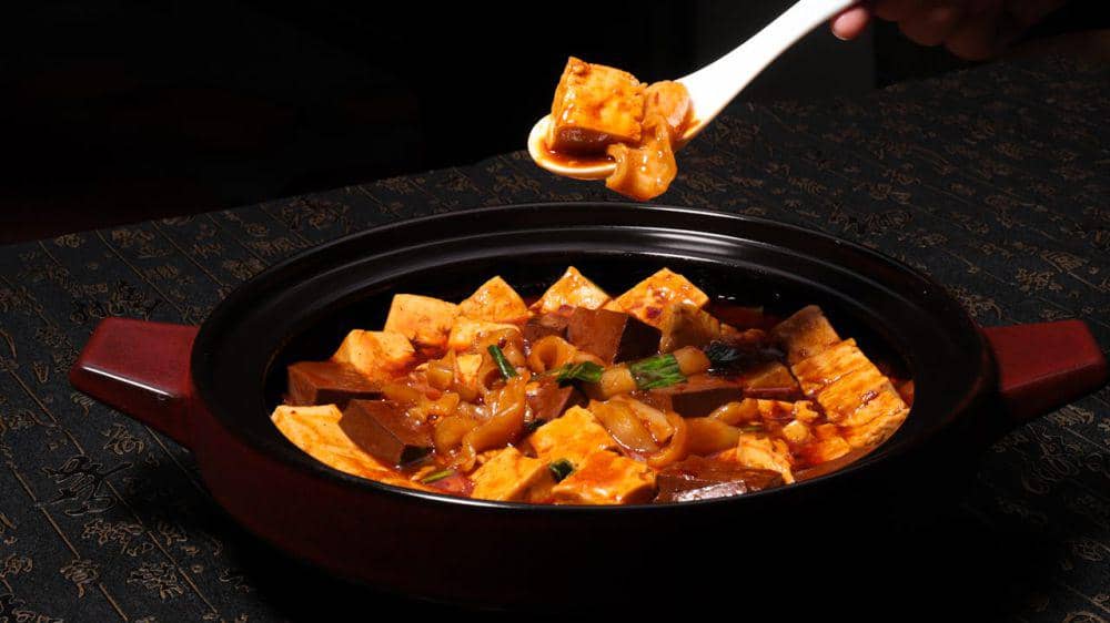 Semangkuk mapo tofu don dengan saus pedas berwarna merah disajikan dalam mangkuk hitam, dengan sendok mengambil potongan tahu.