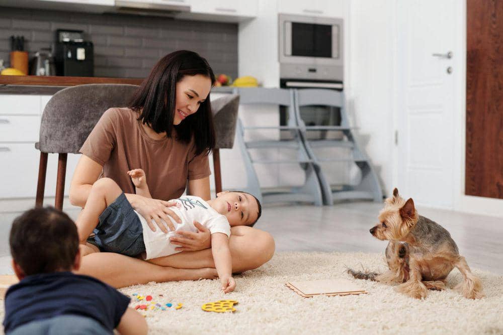 Seorang ibu berdiri mengawasi anak kecil yang bermain bersama seekor anjing di halaman rumah pada siang hari.