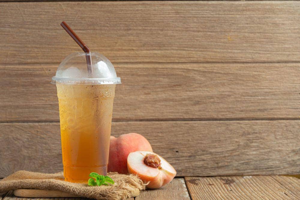 Segelas minuman es teh dengan sedotan cokelat di atas meja kayu, disertai potongan buah persik segar di sampingnya.