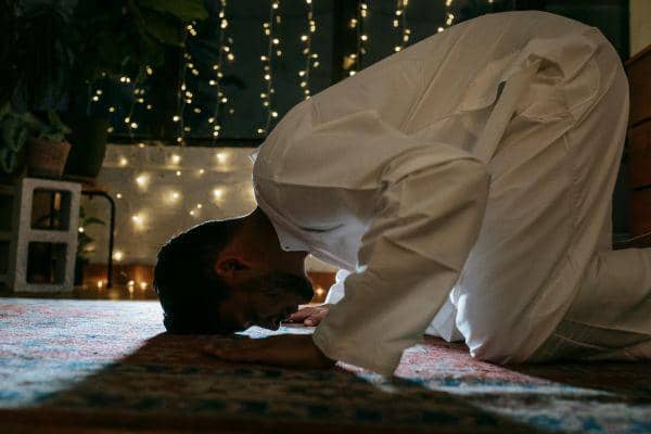 Seorang pria mengenakan pakaian putih sedang sujud saat sholat tahajud di ruangan dengan lampu hias berwarna hangat di latar belakang.