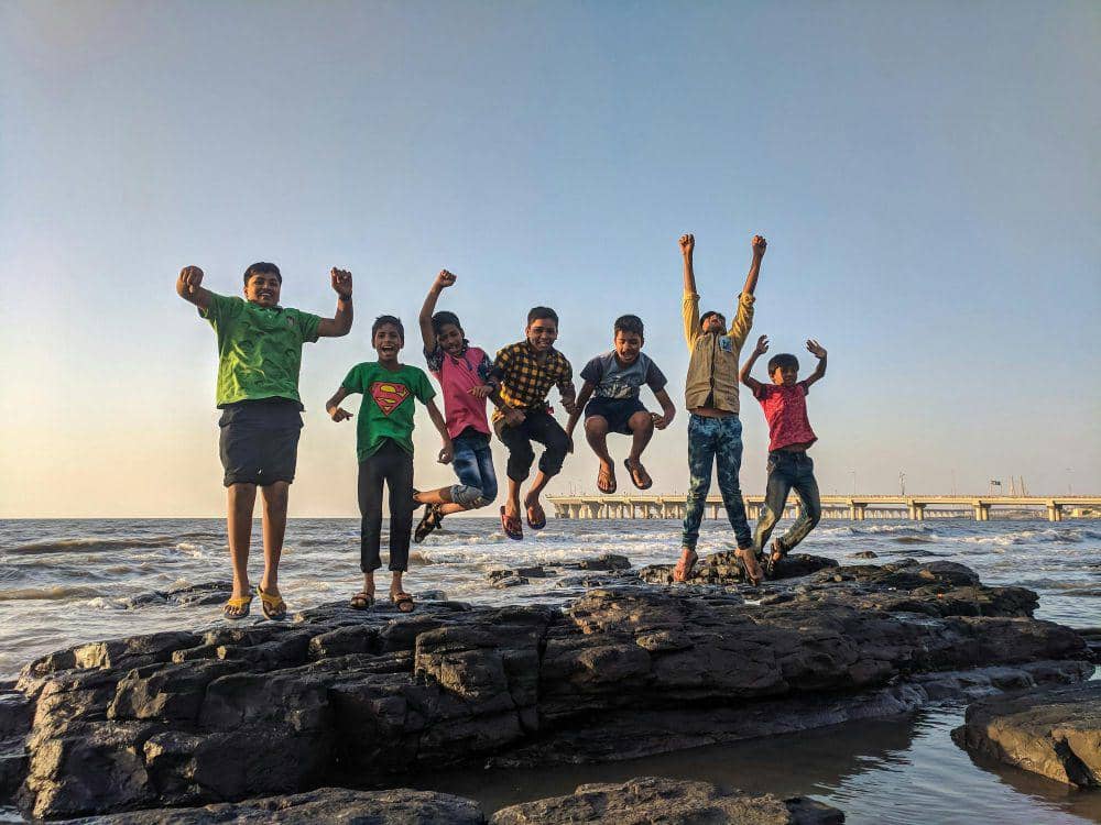 Sekelompok anak-anak melompat gembira di atas batu di tepi pantai dengan latar belakang laut dan langit cerah saat sore hari.