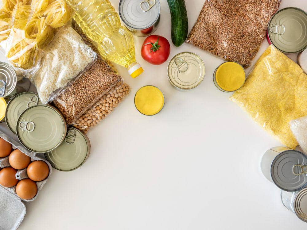 Beragam bahan makanan seperti telur, pasta, minyak, sayuran, dan makanan kaleng tersusun di meja dengan ruang kosong di tengah.