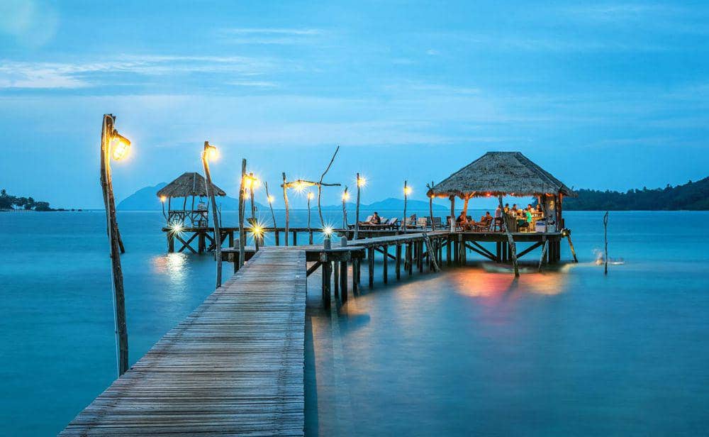 Dermaga kayu dengan lampu-lampu temaram mengarah ke gazebo beratap jerami di atas laut biru saat senja di tepi pantai tropis.