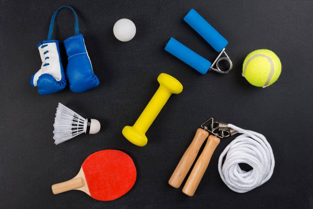 Beragam peralatan olahraga seperti sarung tinju mini, bola tenis, raket pingpong, dumbbell, tali skipping, dan shuttlecock di atas latar hitam.