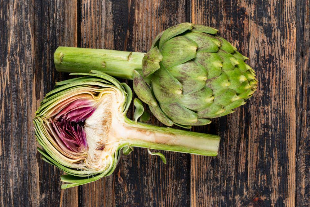 Dua buah artichoke segar di atas permukaan kayu, satu utuh dan satu dibelah memperlihatkan bagian dalam berwarna ungu dan hijau.
