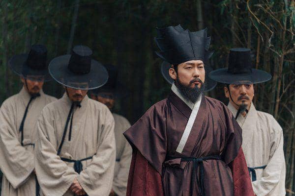 Seorang pria berpakaian tradisional Korea berwarna merah marun berdiri di depan beberapa pria lain yang mengenakan hanbok krem dan topi hitam.
