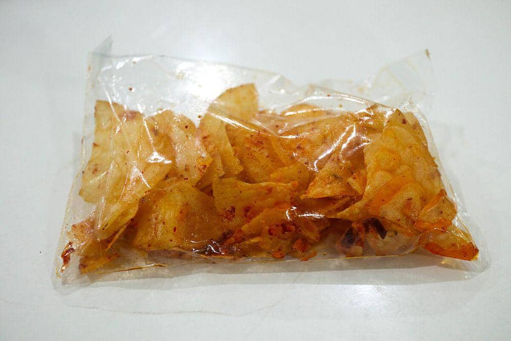 Keripik singkong pedas berwarna keemasan dengan taburan cabai merah dalam kemasan plastik bening di atas permukaan putih.
