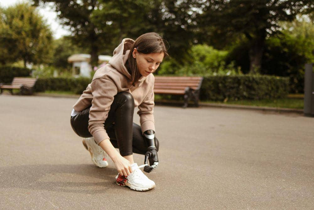 Seorang perempuan mengenakan hoodie dan celana olahraga mengikat tali sepatu sebelum berlari di taman dengan latar pepohonan hijau.