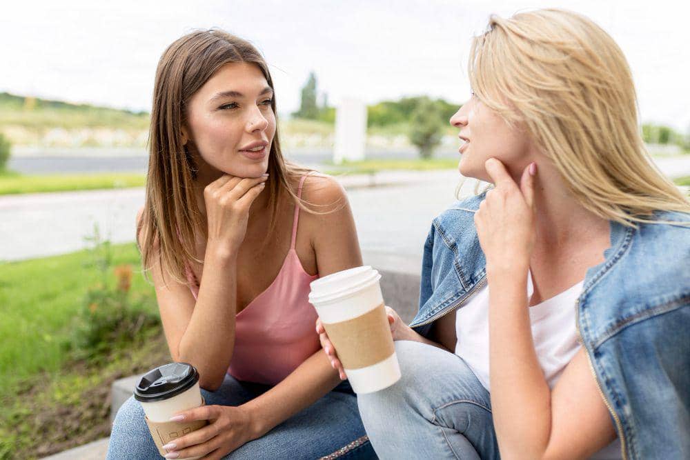 Dua perempuan duduk di luar ruangan sambil mengobrol dan memegang gelas kopi kertas, tampak santai di suasana siang hari.