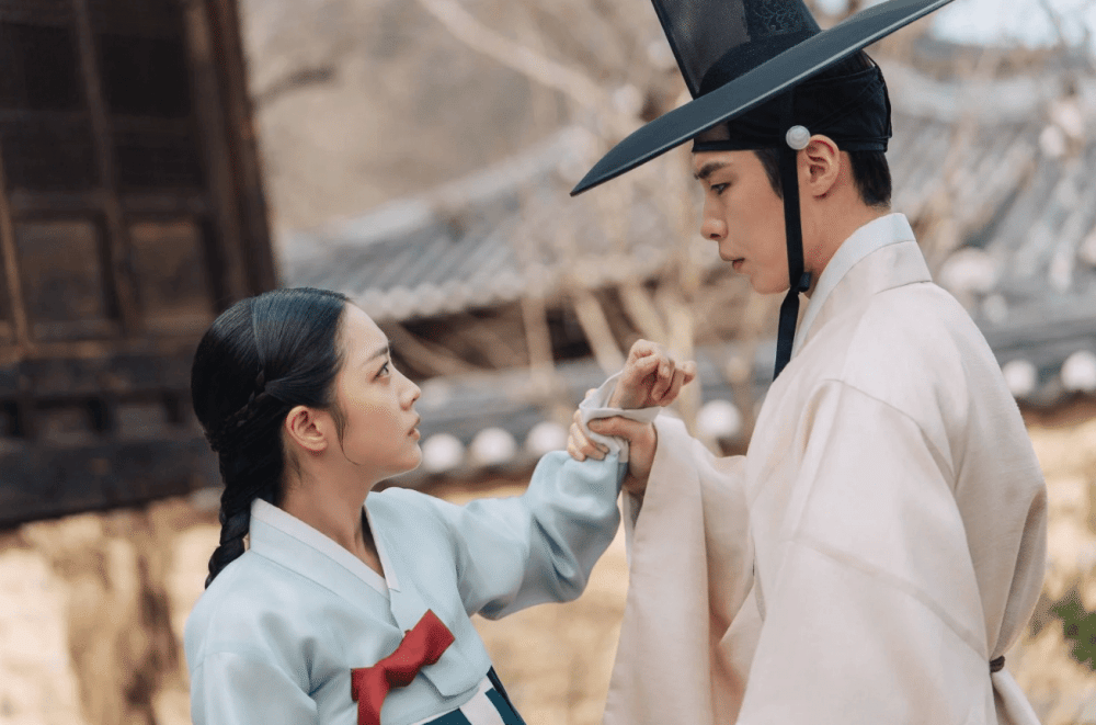 Dua karakter mengenakan hanbok tradisional Korea saling menatap intens di latar bangunan kayu dan halaman kuno.