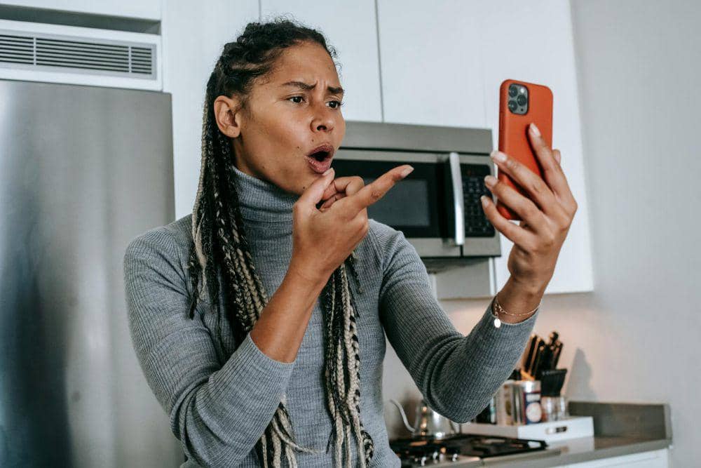 Seorang perempuan mengenakan sweater abu-abu tampak marah sambil memegang ponsel berwarna oranye di dapur modern.